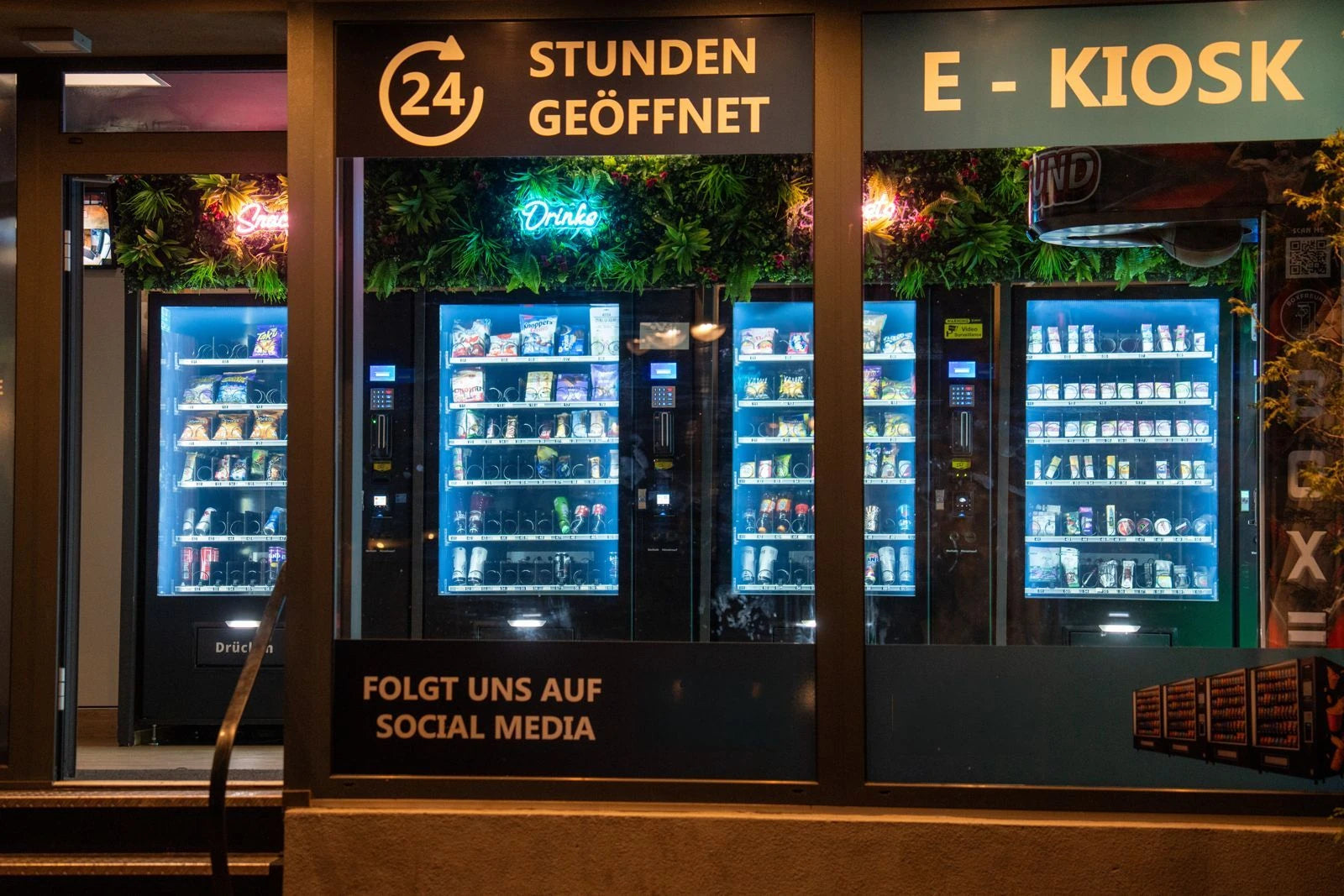 Nachts beleuchteter Automaten-Kiosk mit mehreren Warenautomaten und dem Hinweis „24 Stunden geöffnet"
